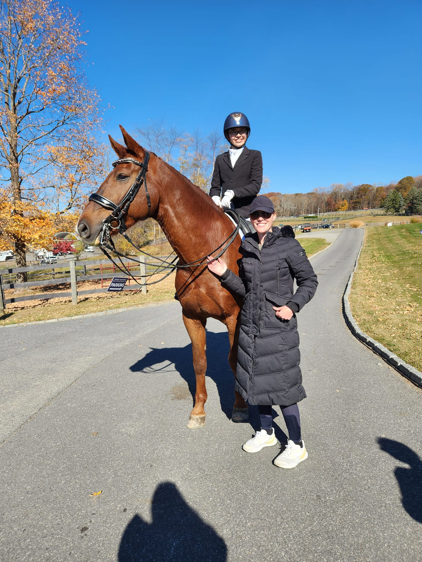 A smiling rider on a chestnut horse with Katie beside them at the farm