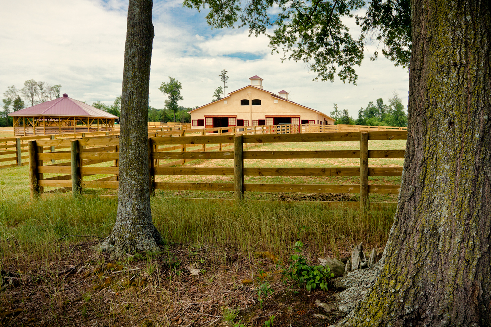 The Equitas grounds—a stable, covered round arena, and post-and-rail fencing under open trees
